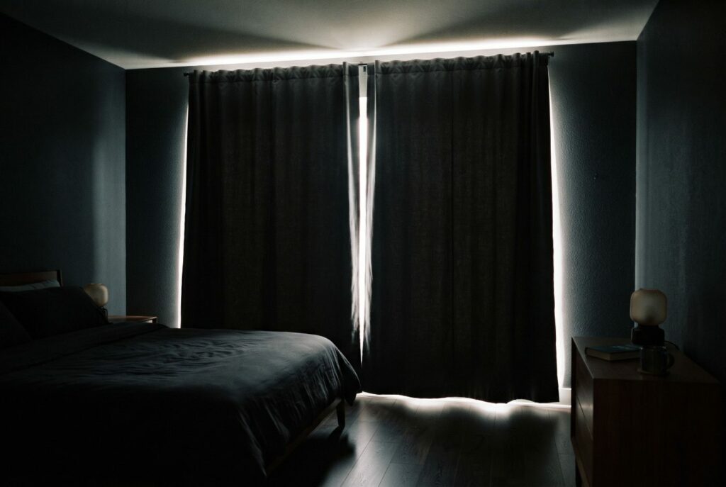 A dark bedroom with closed charcoal curtains and bright daylight leaking over the top, down both sides, through the middle gap and under the hem
