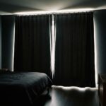 A dark bedroom with closed charcoal curtains and bright daylight leaking over the top, down both sides, through the middle gap and under the hem