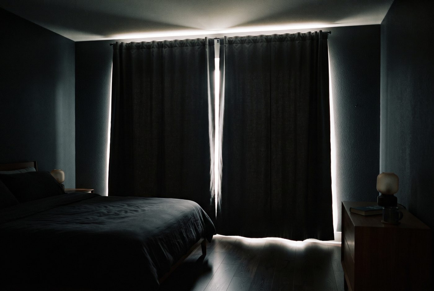 A dark bedroom with closed charcoal curtains and bright daylight leaking over the top, down both sides, through the middle gap and under the hem