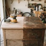 An old wooden dresser in a garage with half of its top sanded to bare wood and a random orbit sander resting on the sanded half