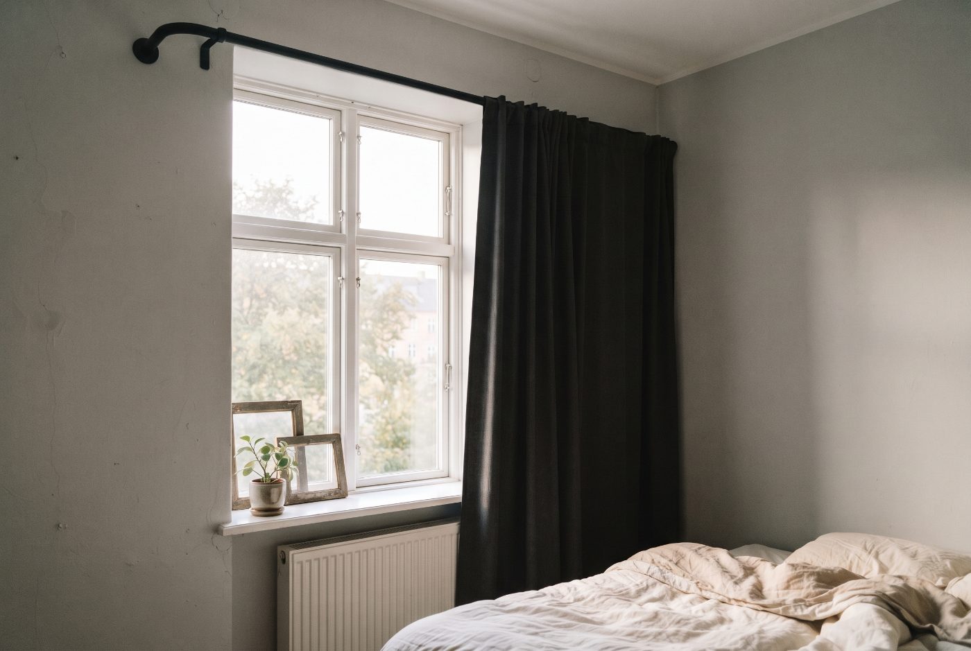 A bedroom window with a dark blackout curtain on a black rod whose curved end turns back into the wall beside the window frame
